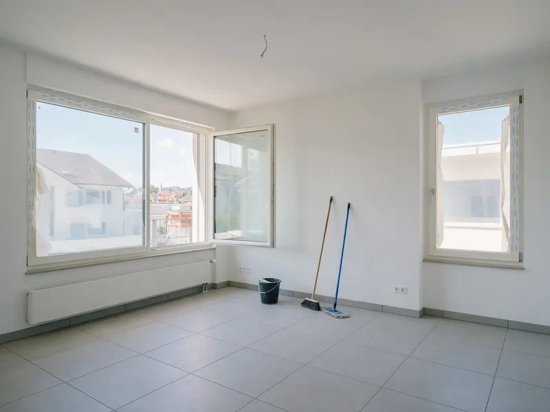 Leere Neubauwohnung mit Plattenboden und grossen Fenstern, Eimer, Besen und Mopp an der Wand